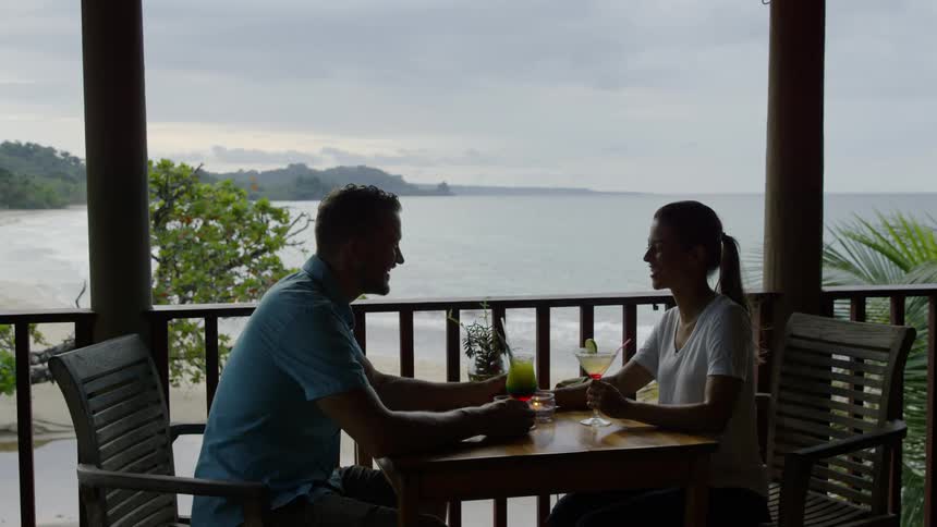 Man and woman are having drinks at table on porch overlooking ocean