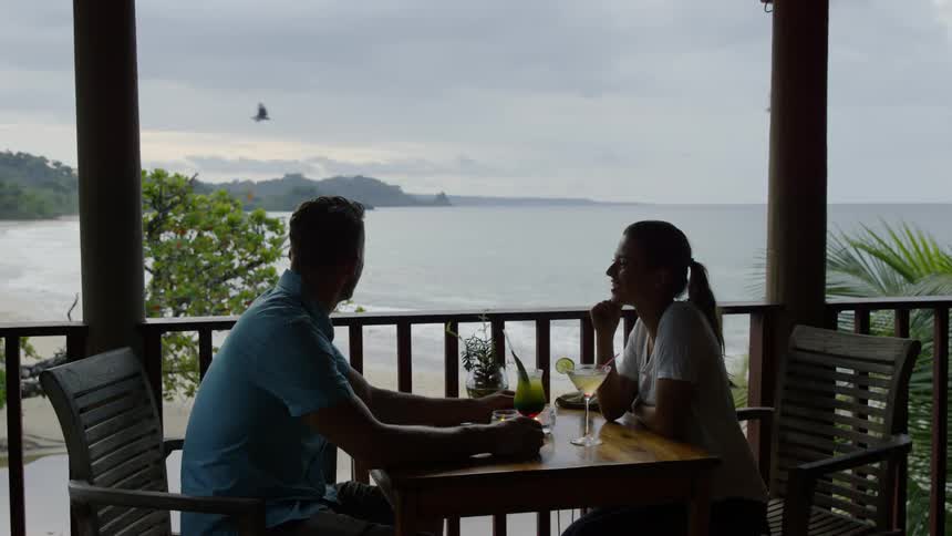Man and woman sit at a table on a balcony overlooking water