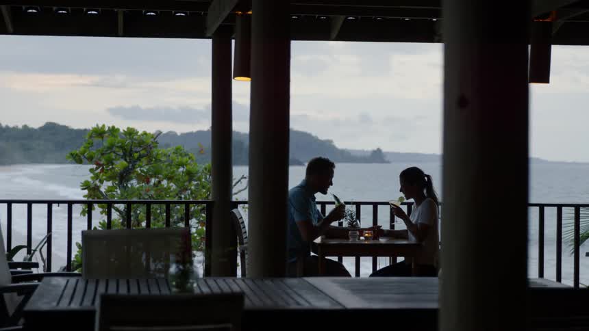 Man and woman drinking at table on porch overlooking the ocean