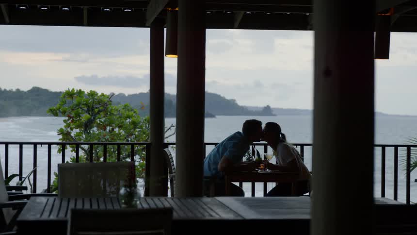 Man and woman sit at a table on a balcony overlooking water