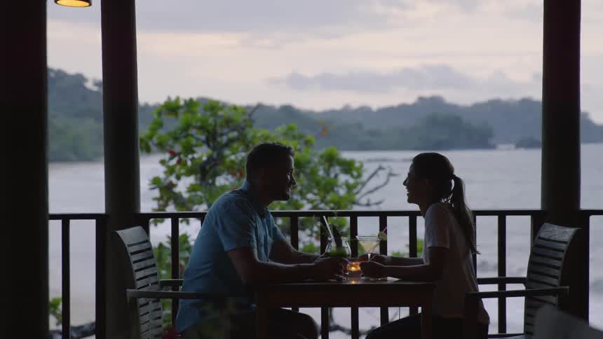Man and woman sit at a table on a balcony overlooking water