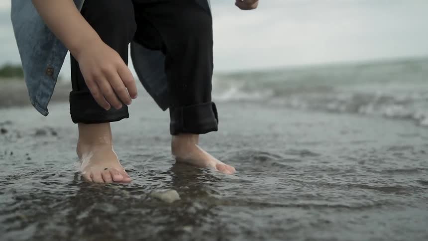 A boy is bending over to pick up a rock from the beach as he wades in the tide