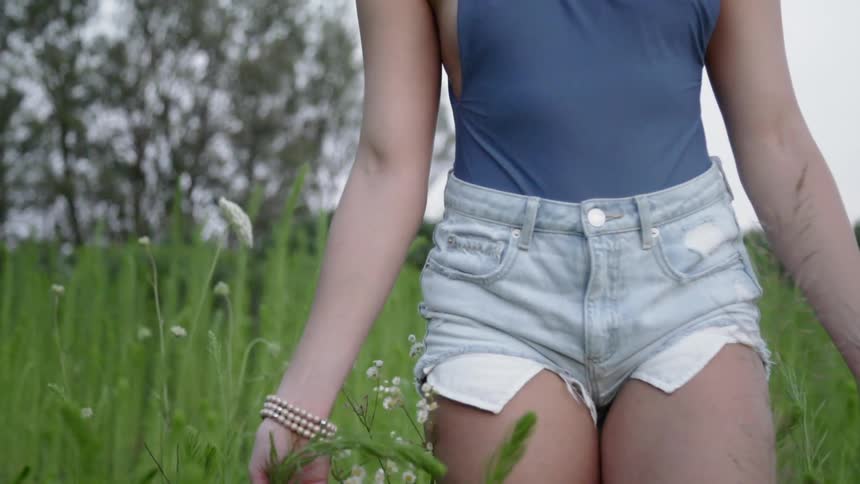 A woman is running her hands through grass and flowers as she walks outside in daylight