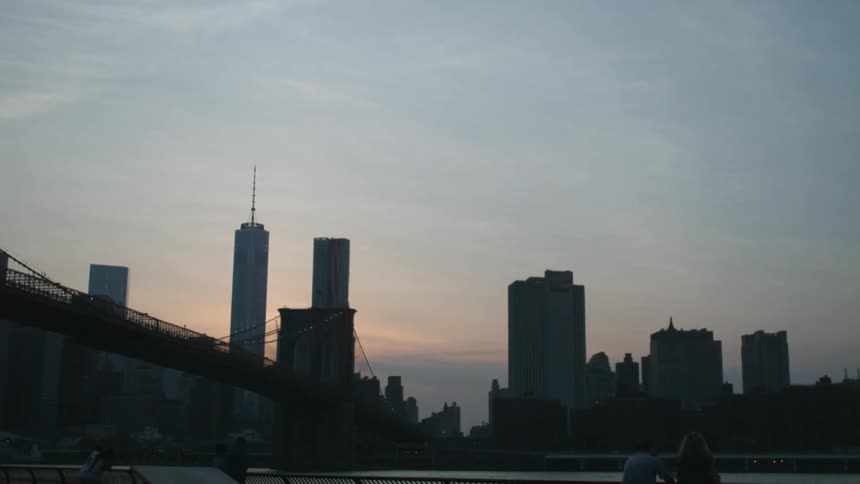 A couple are leaning on a railing viewing a city across a harbor at dusk