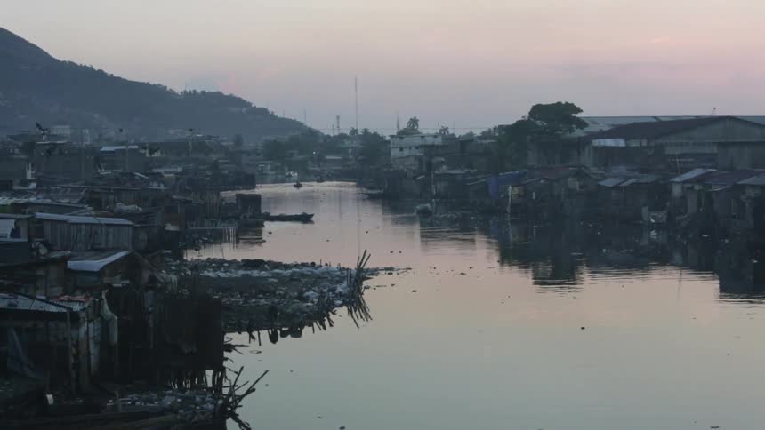 View of rundown shacks that sit along a river bank near a mountain on a cloudy day