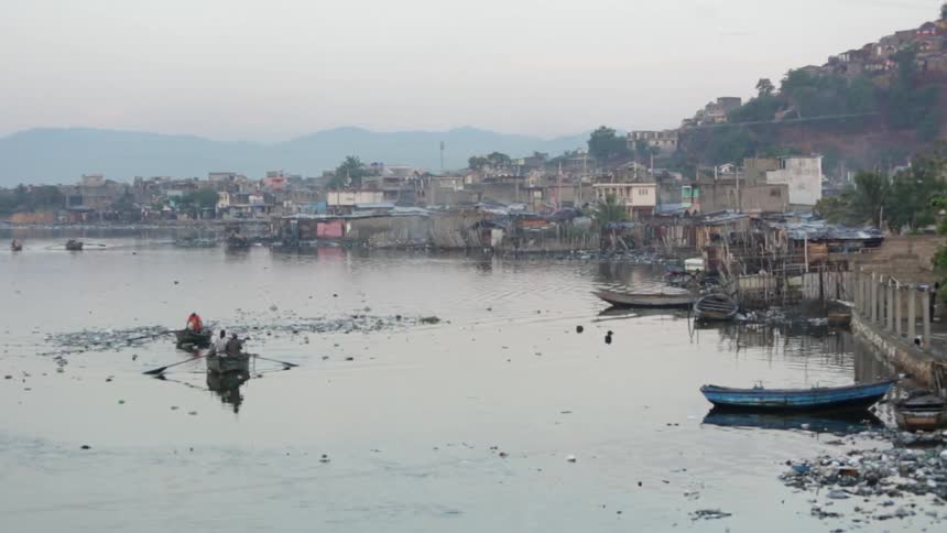 People rowing boats on a body of water located next to homes with mountains in the background