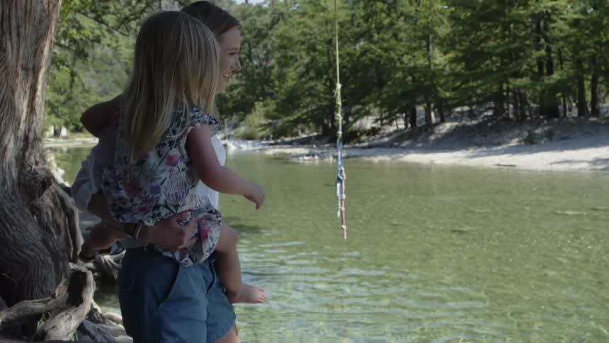 A mother is holding her toddler girl as they are standing watching a river in daylight