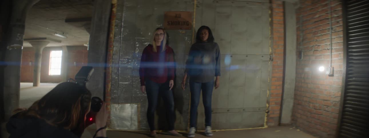 Two women stand in an empty warehouse as a person takes pictures of them
