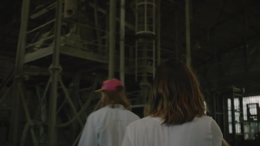 A man and woman walk around an abandoned warehouse with equipment in it