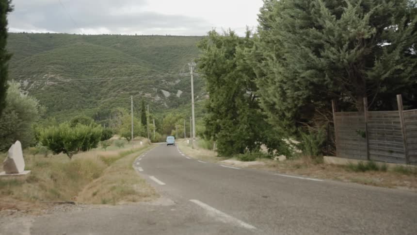 An old blue van drives down a curved road surrounded by trees on a cloudy day