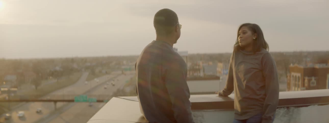 Man and woman stand outside talking on a rooftop with a highway below