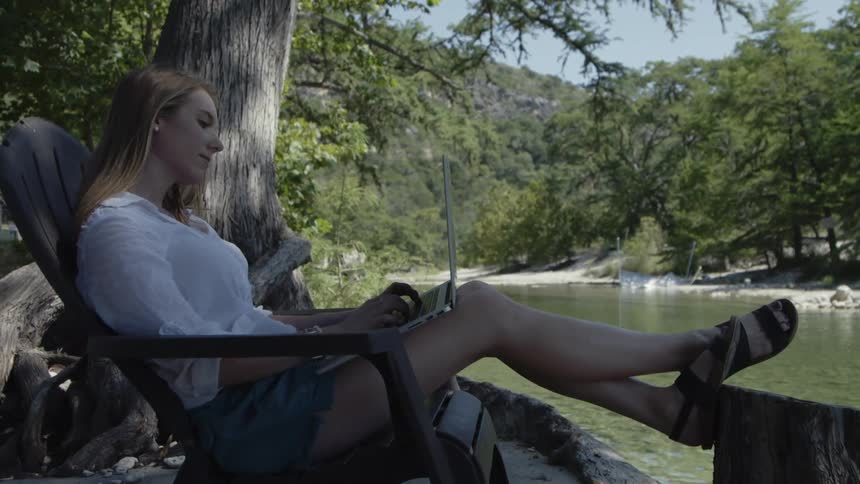 A woman is typing on a laptop near the lake