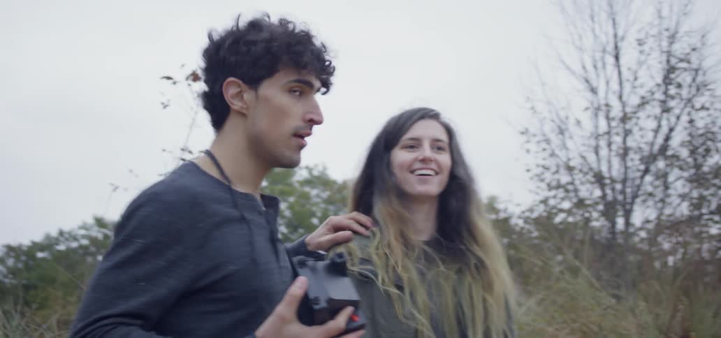 A man is carrying a polaroid camera while walking with a woman in a natural setting