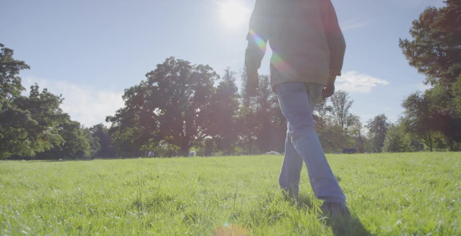 A man walks through a field of green grass with trees and the sun shining in the distance