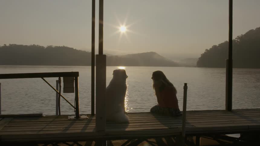 Girl and dog sit on a pier by the water