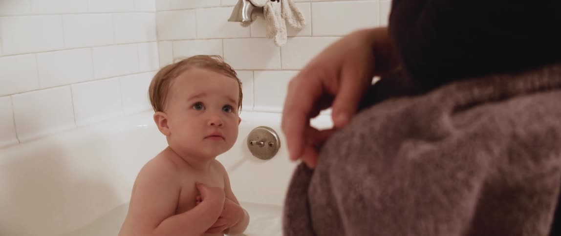 A baby sits and plays in a bathtub while a woman sits and points