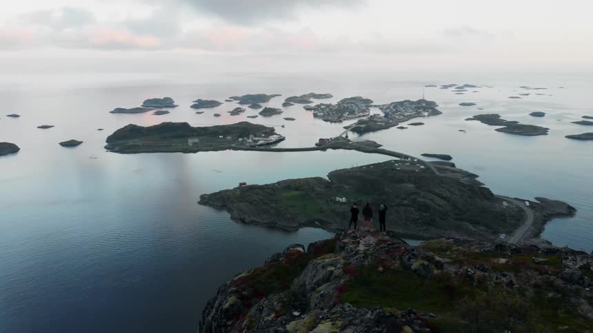Three people stand on a mountain cliff overlooking a large body of water with several small islands