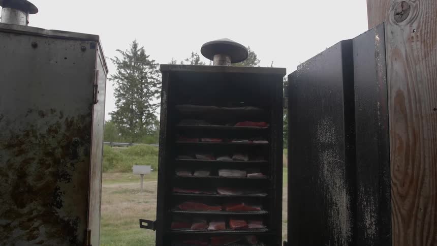 Smoke swirls in front of an outdoor smoker with fish on shelves