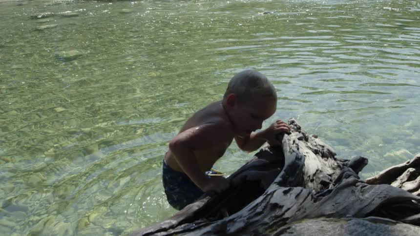 A boy pulls himself up out of a body of water holding onto a piece of wood