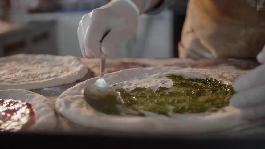 A chef rubs pesto on pizza dough