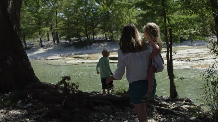 A woman carries a girl towards a boy walking by a stream of water with many trees