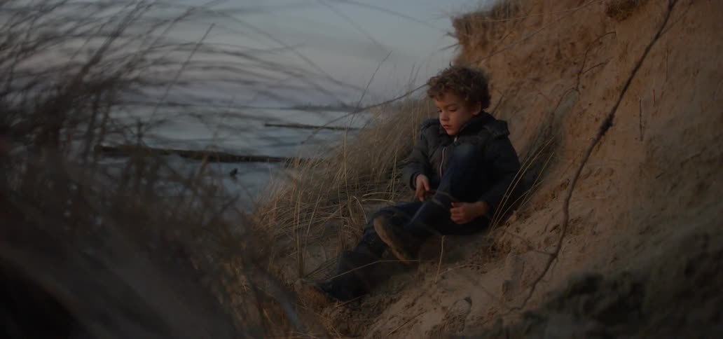 A boy puts on a boot on a beach at sunset