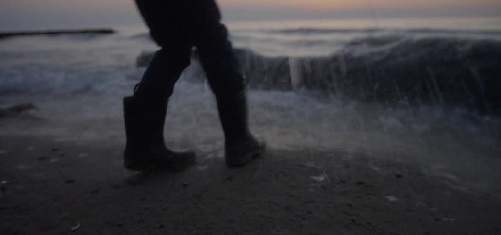 A person wearing rain boots walks near the water on the beach at sunset