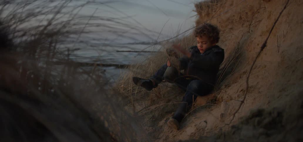 A boy is sitting on a hills shaking the sand out of his boots at dusk