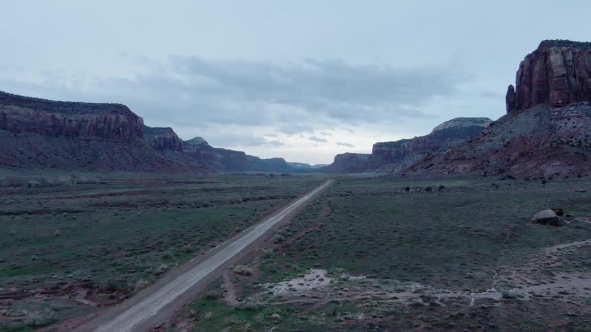 A road runs through a large grassy area surrounded by mountains with clouds in the sky