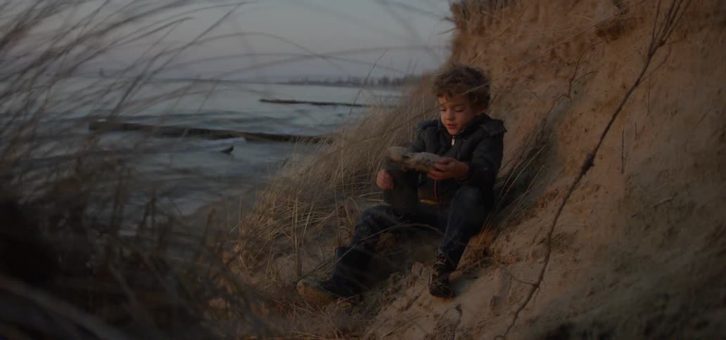 Young boy sits on a hill as he dumps sand from his boot