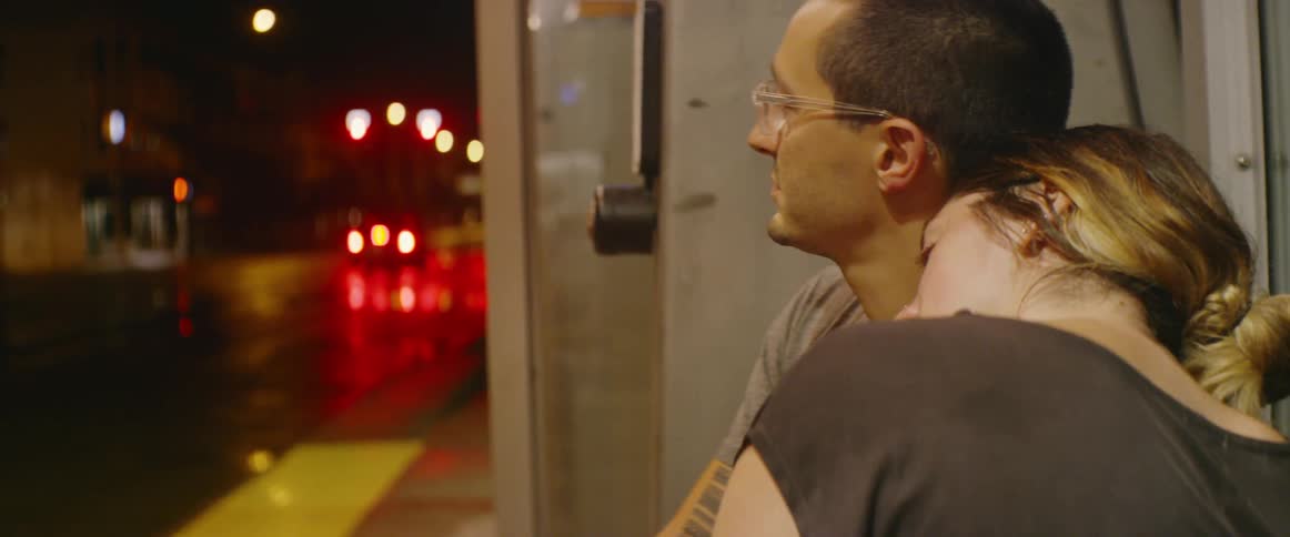 A woman leans against a man's shoulder as they sit at a bus stop at night