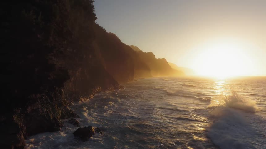 The waves crash against the tall cliff at sunset