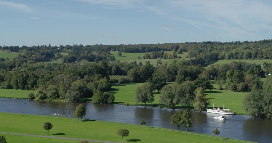 A small boat travels past a larger one down a stream surrounded by grass and trees on a sunny day