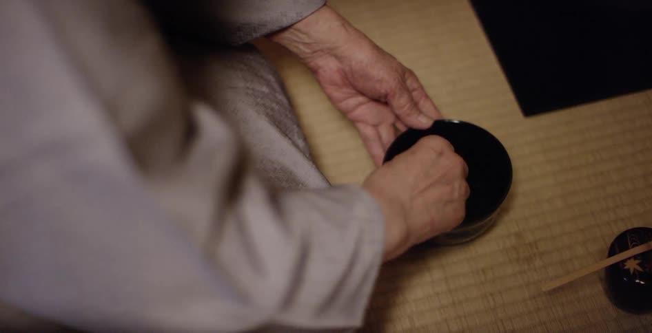 An elderly person sits at a table and scrubs a small bowl