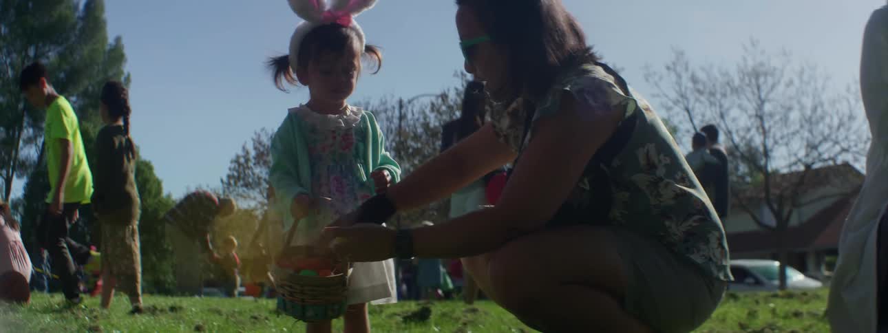 A woman crouches down next to a young girl wearing bunny ears and holding an easter basket at an egg hunt