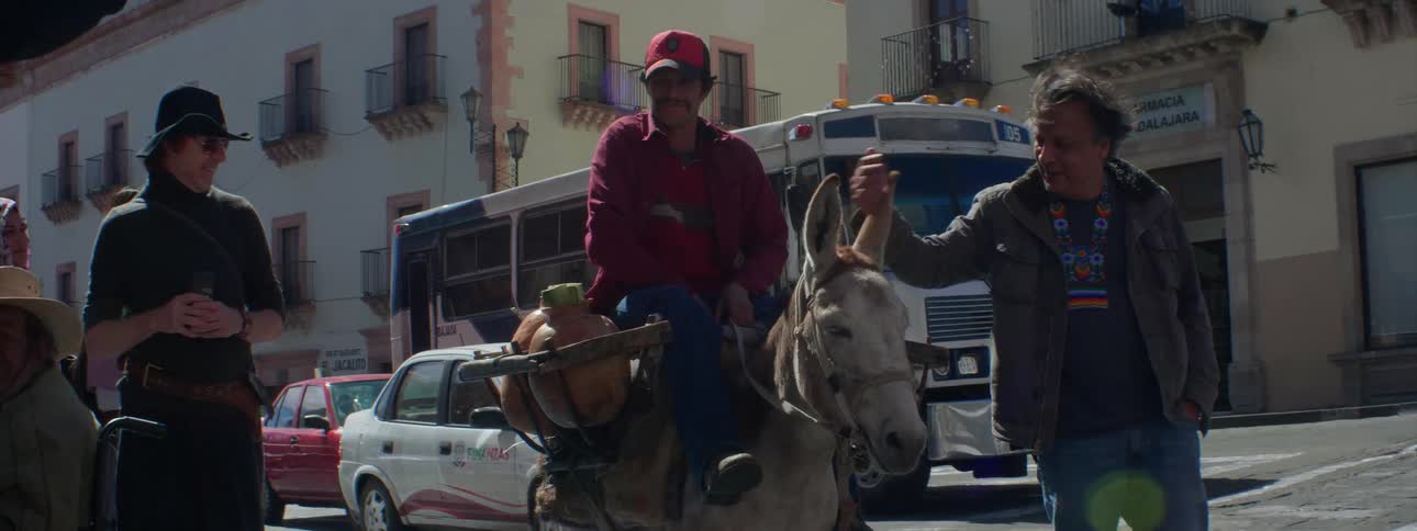 A man rides a donkey in a city street as another pets the donkey on the head