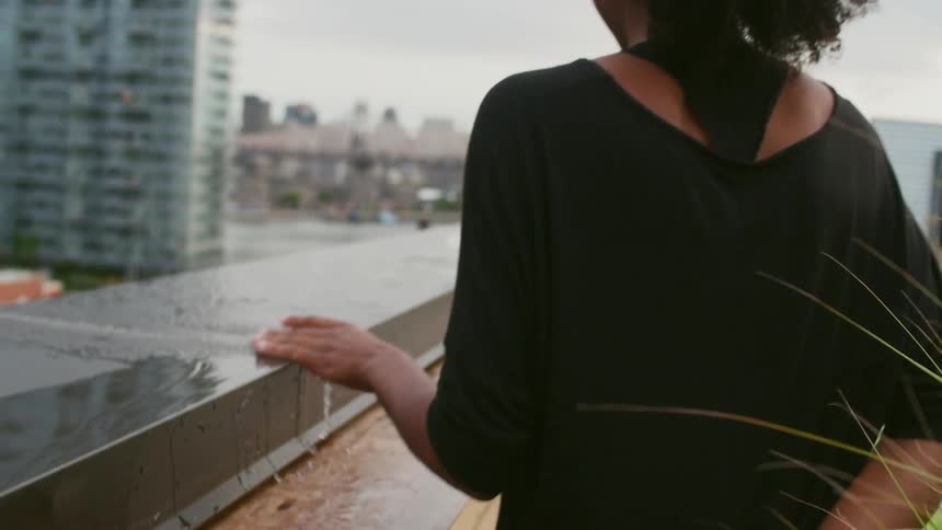A young black woman running her hand along the top of a wet wall as she walks and smiles over her shoulder in an urban area
