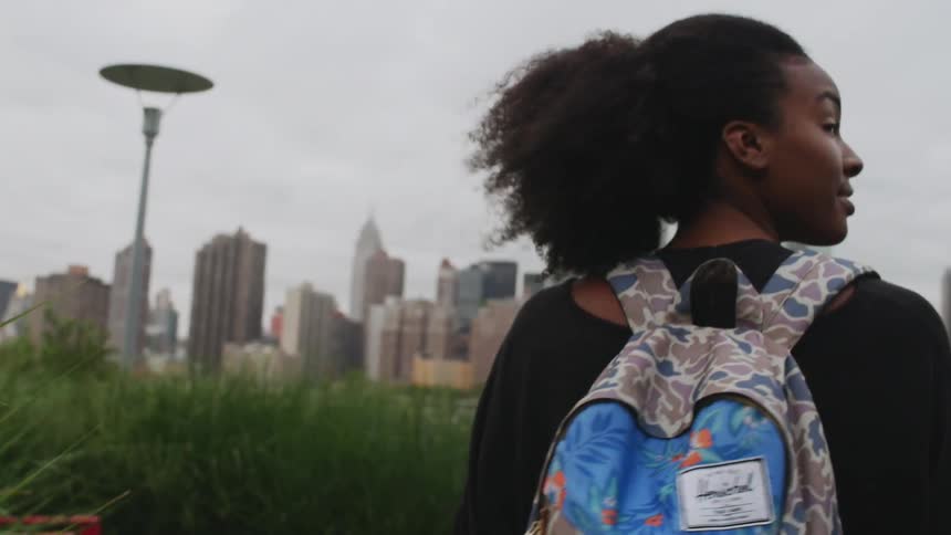 A girl wearing a backpack walking through grass towards a city