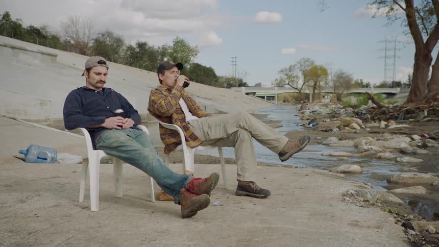 Two men drink beer in lawn chairs next to a creek