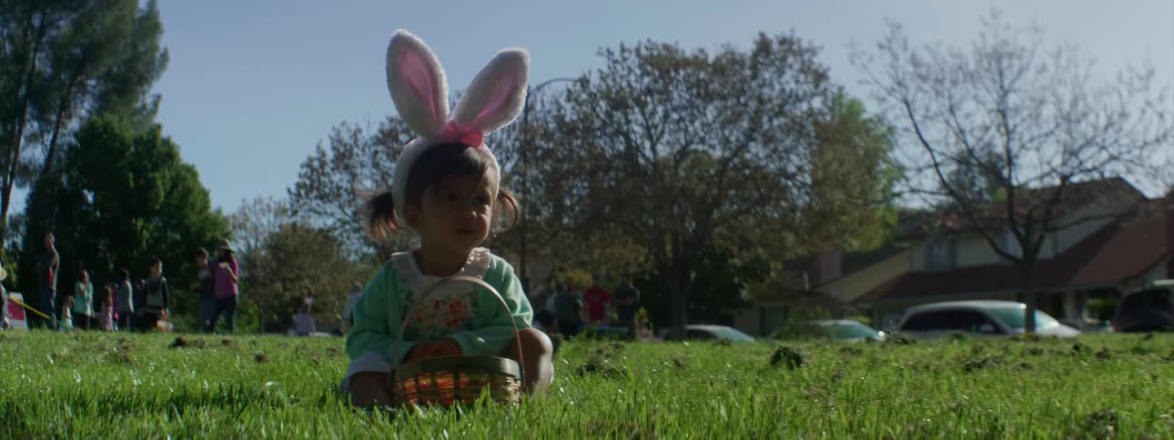 Young girl wearing costume bunny ears sitting with her basket in a field as others walk around behind her on a clear day