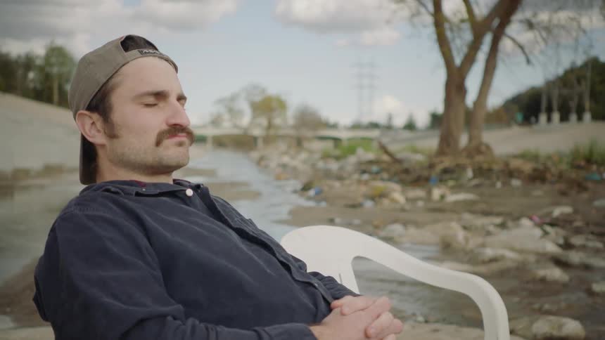 Man sits in a chair and puts a can down on the ground as another man stalks up to hit him with a water jug
