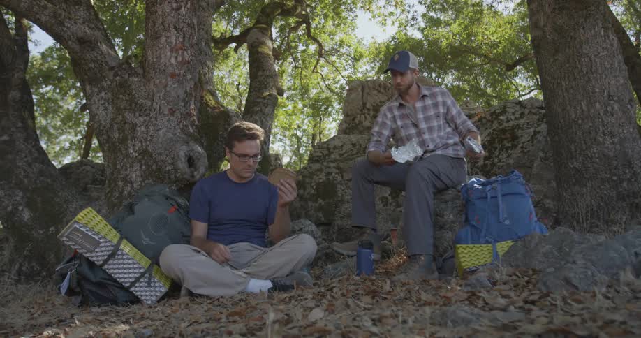 Two men sit in the woods on a sunny day eating sandwiches