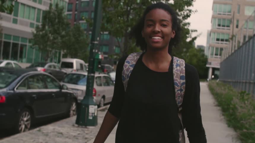 A smiling person in a black shirt walks down the sidewalk