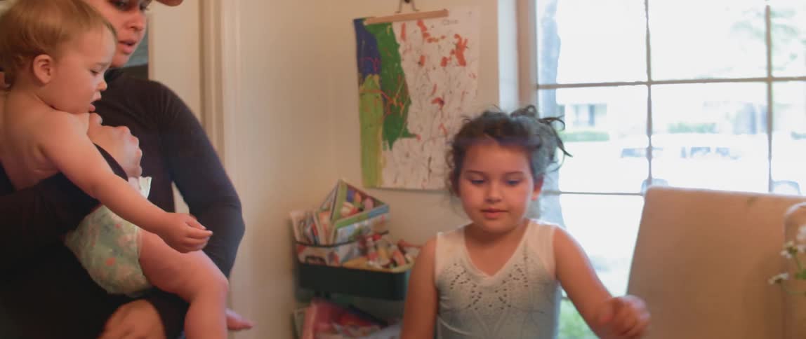 A woman holds a baby while she talks to a little girl and they look at her artwork at a dining room table