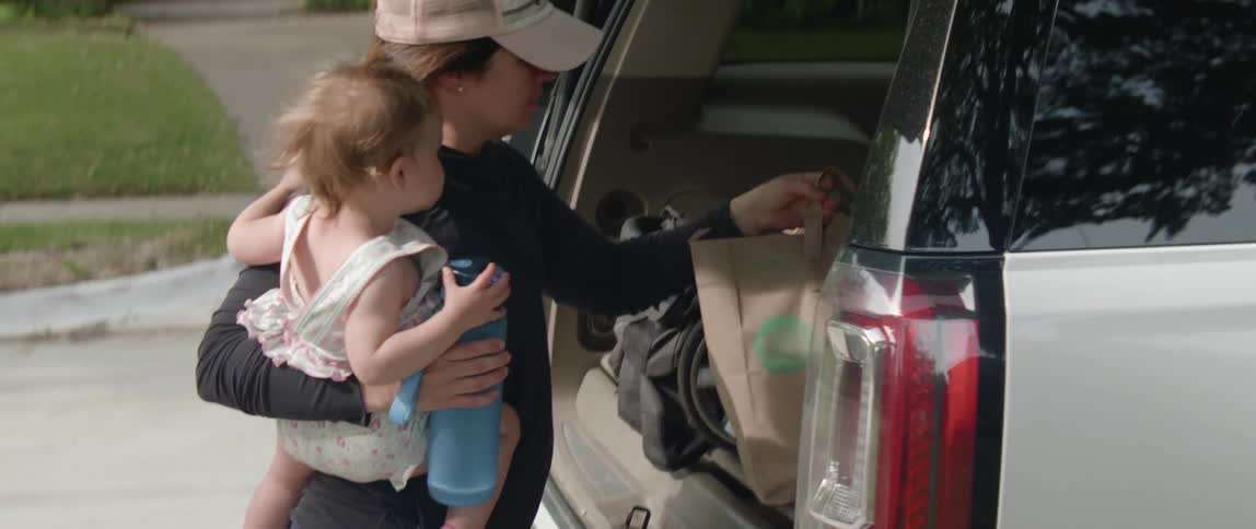 A woman holds a baby as she unloads groceries from the trunk of a car