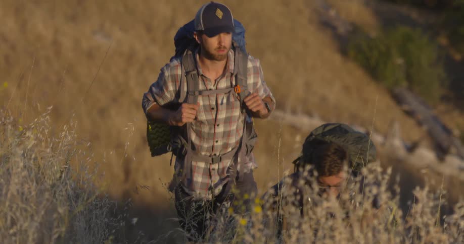 Two men wearing backpacks walk up a hill under a sunny sky