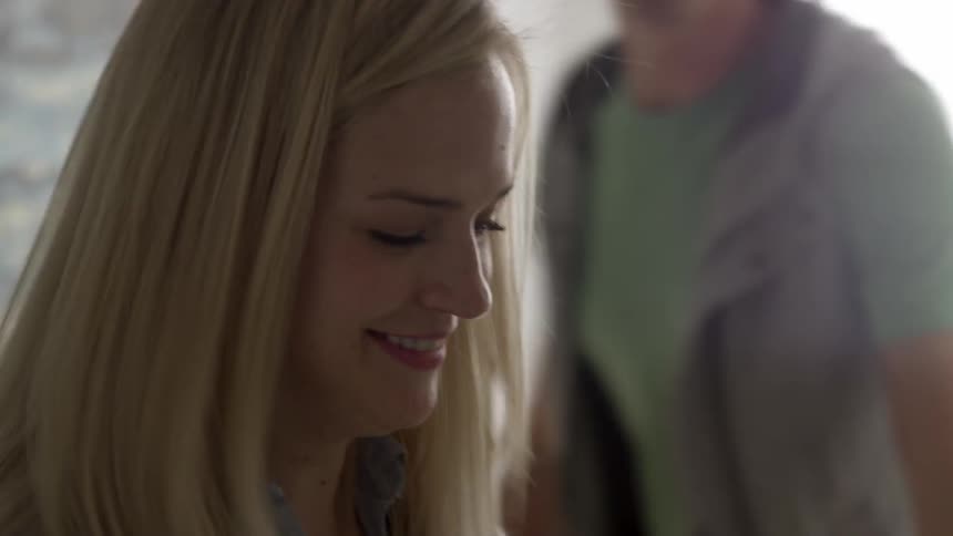 A young, blonde woman looking down at something and smiling before a man enters the area and she turns to kiss him