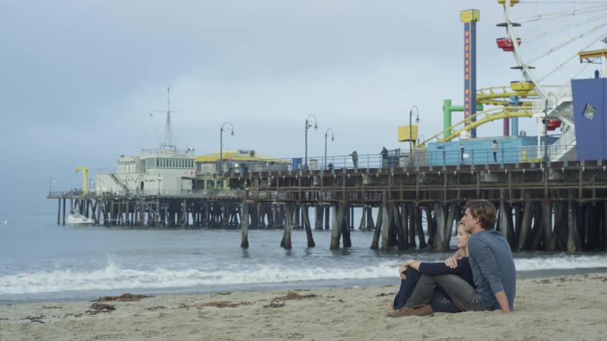 A man and woman sitting on a beach next to an amusement park