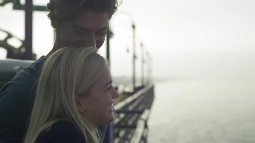 A blonde man putting his arm around a blonde woman as they talk and laugh while looking out over the water