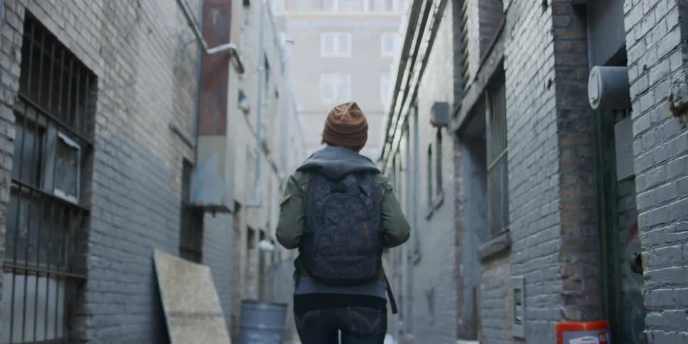 A woman wearing a backpack walks down an alley and looks back